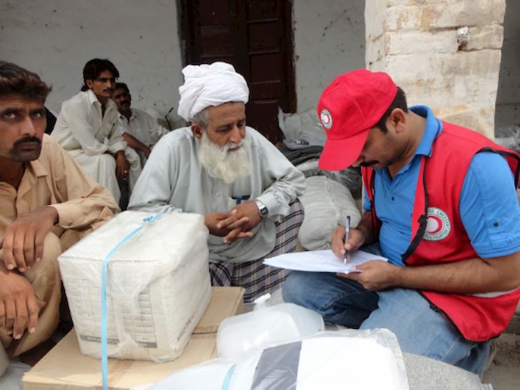 A Pakistani elder wearing a turban receives a relief package from a Red Cross worker who is filling out a form