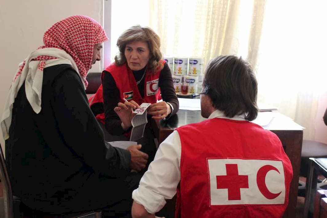 Two Red Cross Red Crescent workers - a man and a woman - explain some information on a leaflet to a man in a red headscarf.